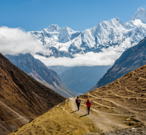 Zwei Wandernde auf einem Bergpfad zwischen steilen T&auml;lern, vor schneebedeckten Himalaya-Gipfeln.