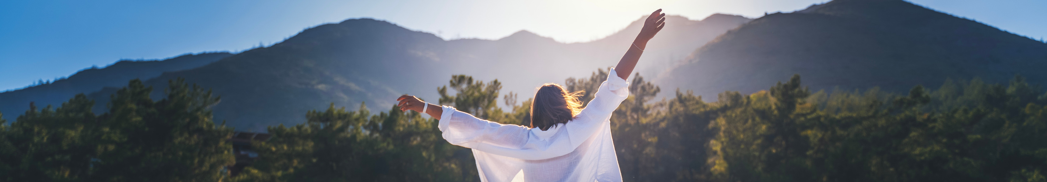 Frau mit ausgebreiteten Armen vor bewaldeten Bergen bei Sonnenaufgang 