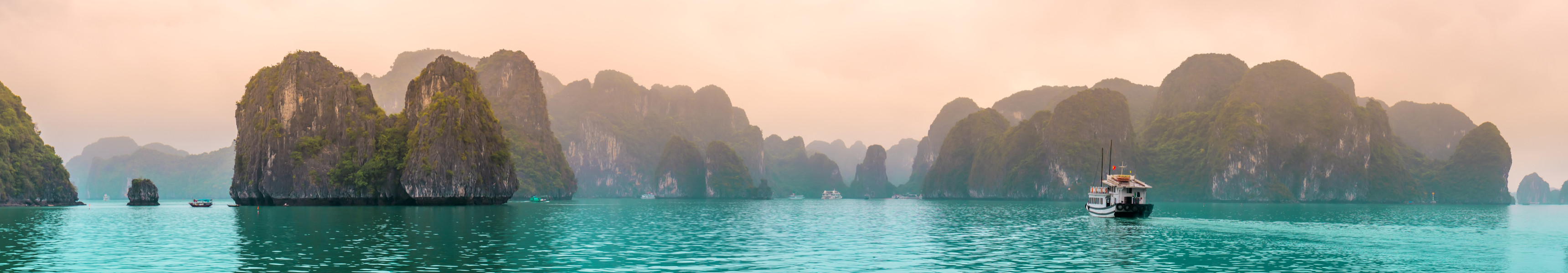 Panorama der Halong-Bucht in Vietnam mit t&uuml;rkisfarbenem Wasser, Kalksteinfelsen und einem Boot im Vordergrund bei nebliger Abendstimmung
