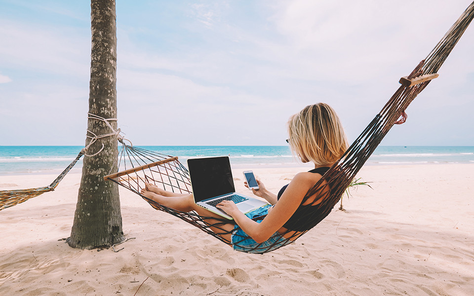 Frau im Strandurlaub arbeitet digital mit Laptop und Smartphone in H&auml;ngematte.