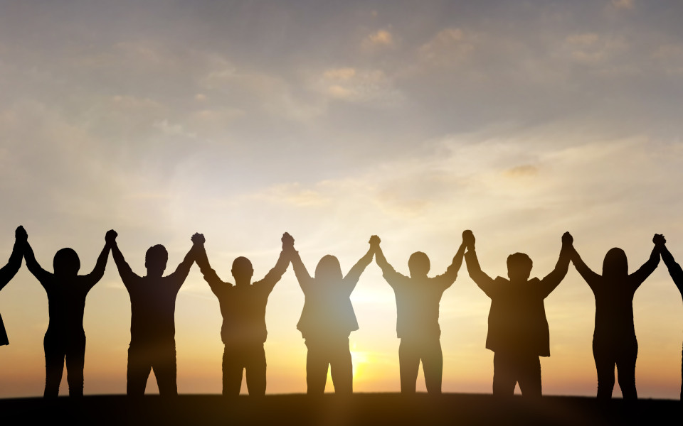 Silhouetten einer Gruppe von Menschen, die bei Sonnenuntergang nebeneinander stehen und die H&auml;nde in die H&ouml;he halten.